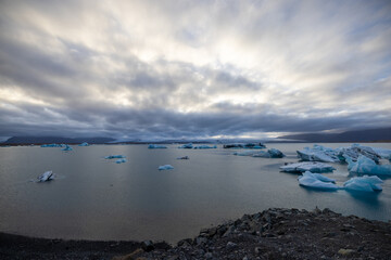 Icebergs peacefully float on the liquid surface of the lake, under a cloudy sky with cumulus clouds, creating a serene natural landscape. Jokulsarlon Glacier Lagoon, Iceland