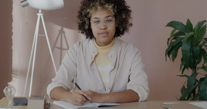 Slow motion portrait of good-looking African American lady entrepreneur writing in notebook doing business planning turning to camera smiling in office. Workplace and businesswoman concept.