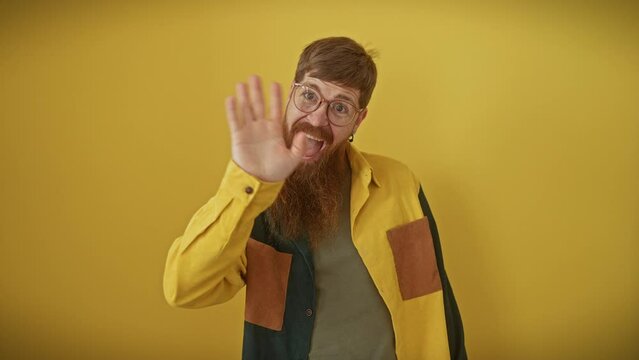 Joyful young redhead man standing over an isolated yellow background, smiling and waving hello. confident guy in glasses and shirt with a friendly, welcoming gesture.