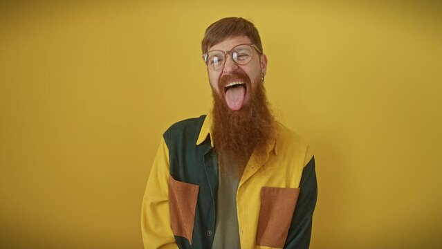 Cheeky young redhead guy sticking out tongue with a funny expression, happily standing over isolated yellow background wearing glasses and shirt.