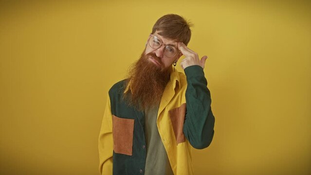Depressed young redhead guy, glasses on, shirt-clad, standing over yellow, staged isolation, reenacts suicide by mock-shooting himself, hands gesturing guns at his head.