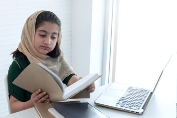 Young Muslim girl intent on learning with exercise book and laptop on desk