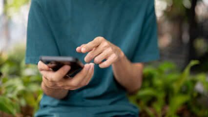 A cropped image of a man in casual wear using his smartphone outdoors, sitting on a bench in a park.