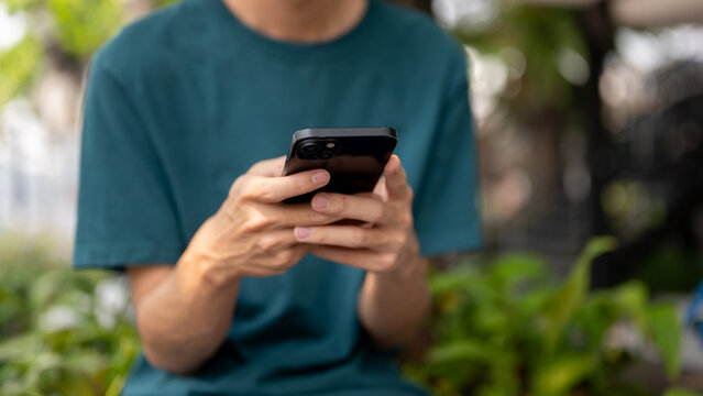 A cropped image of a man in casual wear using his smartphone outdoors, sitting on a bench in a park.