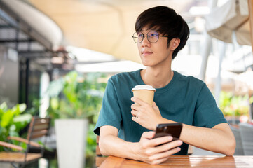 A man holding a coffee cup and a smartphone, looking away, sitting at an outdoor table of a cafe.