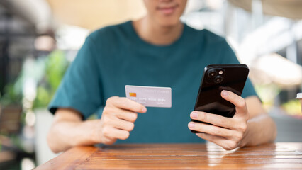 A young Asian man holding a credit card and a smartphone, sitting at a wooden table outdoors.