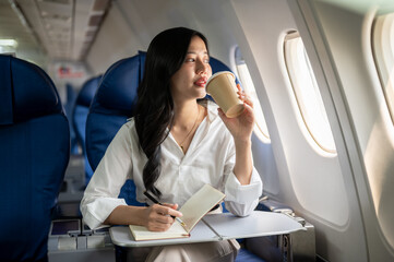 An elegant Asian businesswoman is sipping coffee while looking at the view outside the plane window.
