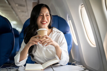 An elegant Asian businesswoman is sipping coffee while looking at the view outside the plane window.