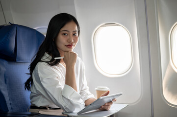 A confident Asian businesswoman is sitting at a window seat on a plane with her digital tablet.