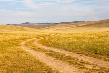 Dirt road on sandy soil in the steppe with green and yellow grass on the meadows of Olkhon Island at sunset. The coast of the Siberian Lake Baikal. Summer landscape. Travel and long journey concept