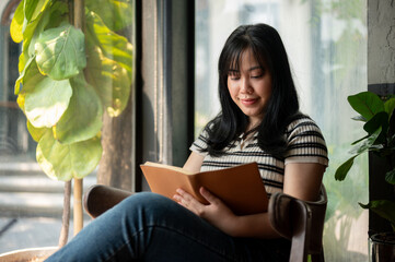 A calm, relaxed Asian woman is enjoying reading a book in a beautiful green cafe.