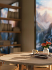 A wooden table with a flower vase, a coffee cup, and books in a room with a mountains view.