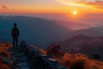 Foto op Plexiglas A man stands on the summit of a mountain as the sun sets in the background © MastersedZ