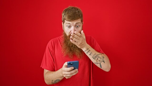 Cool, handsome redhead man stumbles upon surprise message on smartphone over isolated red background