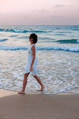 Happy woman in a white dress enjoys relaxing on the beach, walking along the sandy shore at sunset. - 776952251