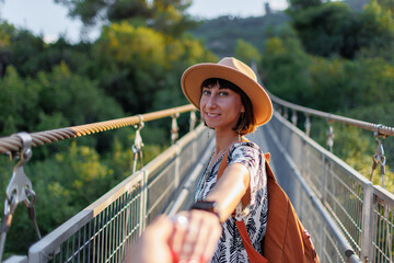 Fall season outdoor portrait woman walking on hanging bridge over river, girl turning around and giving hand to camera like follow me - 776960869