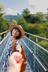 Girl with a backpack. outdoor portrait of a young woman walking on a suspension bridge over a river, the girl turns and extends her hand to the camera as if she is following me. - 776961245