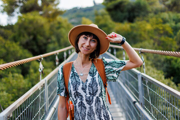 Girl with a backpack. outdoor portrait of a young woman walking on a suspension bridge over a river. . - 776961814
