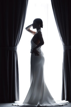 Portrait Bride woman in Silhouette Against Window Light in Elegant Wedding Dress, modern fashion photo, back view