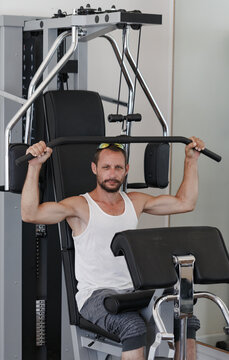 European man using a gym machine pulling down a bar attached to weights wearing a tank top and grey shorts showcasing muscular arms. The machine is black and silver located in gym with light walls