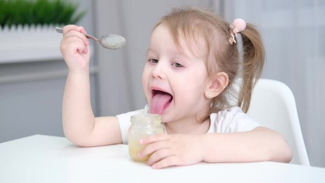 cute little caucasian baby girl eating shredded baby food vegetable puree by herself with a spoon. child eating lunch smiling and looking at camera.