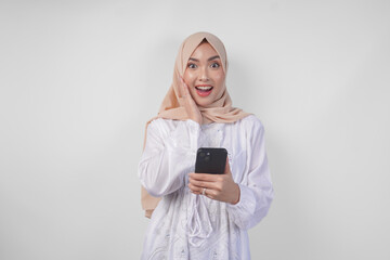 Fototapeta premium Excited Asian woman wearing hijab and white dress smiling happily while holding smartphone, standing over isolated white background. Ramadan and Eid Mubarak concept
