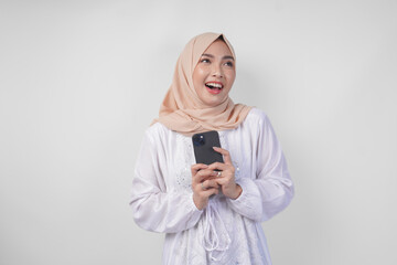 Thoughtful young Asian Muslim woman wearing white dress and hijab holding phone while thinking and finding idea with happy expression, isolated on white background. Ramadan and Eid Mubarak concept