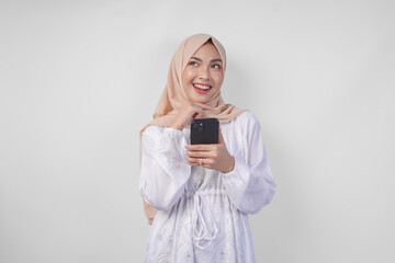 Thoughtful young Asian Muslim woman wearing white dress and hijab holding phone while thinking and...