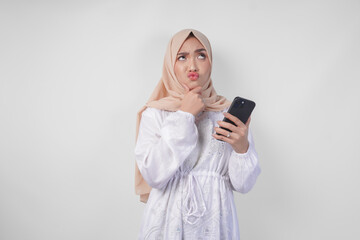 Fototapeta premium Thoughtful young Asian Muslim woman wearing white dress and hijab, using smartphone while holding her chin and thinking with serious expression over isolated white background