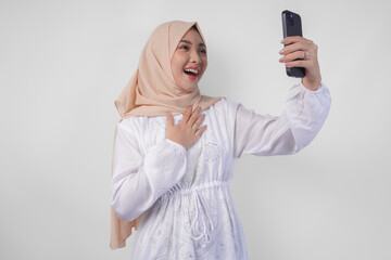 Portrait of Asian Muslim woman wearing white dress and hijab holding her smartphone to do a video call to family or friend, isolated by white background. Ramadhan and Eid Mubarak concept