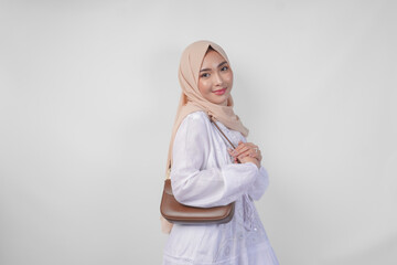 Fototapeta premium Stylish young Asian Muslim woman wearing white dress and hijab with brown sling bag smiling happily over isolated white background. Ramadhan fashion concept