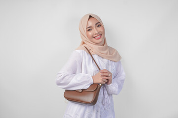 Stylish young Asian Muslim woman wearing white dress and hijab with brown sling bag smiling happily...