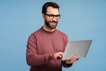 Young man, bearded freelancer, hipster wearing eyeglasses holding laptop, typing on keyboard