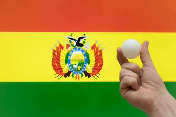 The hand holds a table tennis white ball in front of Bolivia flag.