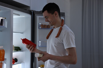 Happy man with sausages and sauces near refrigerator in kitchen at night