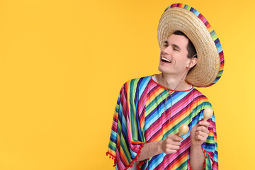 Young man in Mexican sombrero hat and poncho with maracas on yellow background. Space for text - obrazy, fototapety, plakaty