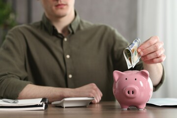 Financial savings. Man putting dollar banknote into piggy bank at wooden table, closeup