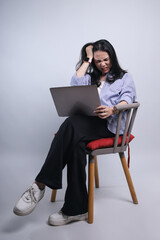 Full length view of Asian woman sitting  on a chair with laptop on her laps and holding her head suffering from pain or headache 