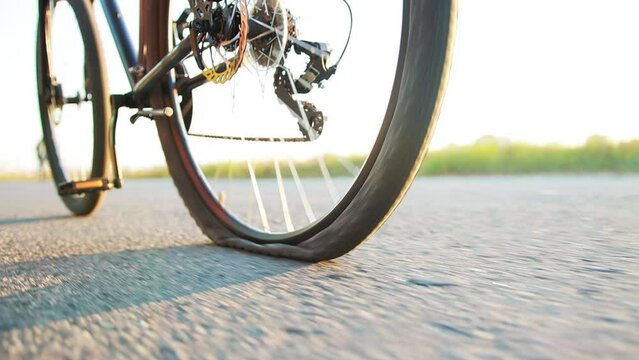 A man rolls a bicycle with a punctured wheel on the asphalt against the backdrop of the summer sun, close-up. Slow motion