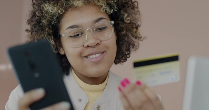 Happy African American lady shopping with smartphone buying in online store paying with credit card smiling at home. Gadget and consumerism concept.