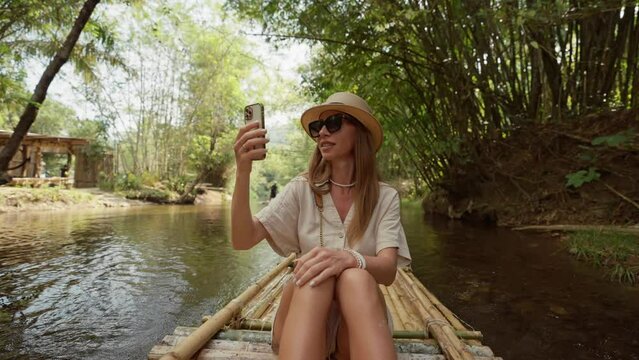 Female tourist floats on bamboo raft. Woman tourist enjoying the bamboo rafting on the river, films beautiful nature landscape on smartphone. Vacation, tropical tourism, jungle, adventure concept.