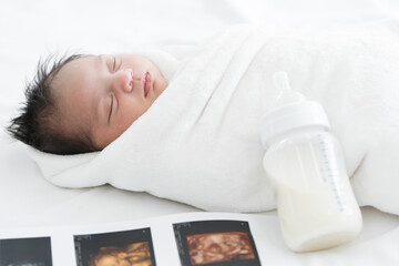 Portrait of Asian Australian newborn baby less than one month old wrapped with cotton towel sleeping on bed with fetal ultrasound image for pregnancy report and milk bottle. New born kid concept