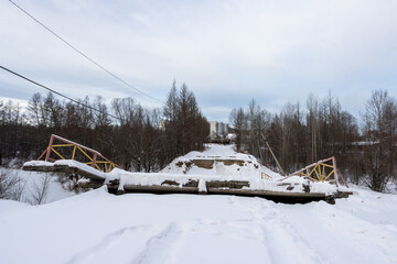 View of the collapsed road bridge. Collapsed bridge over a small river. Fallen damaged bridge. Consequences of an emergency. Cold winter weather. A lot of snow. Residential buildings in the distance.