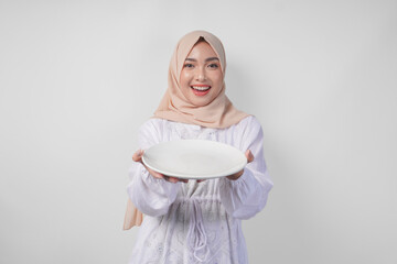 Beautiful young Asian Muslim woman in hijab presenting an empty plate with copy space over it,...
