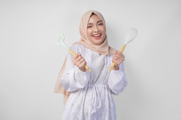 Portrait of a happy Asian Muslim woman in veil hijab holding spatula and kitchen cooking utensils...