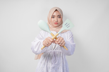 Displeased Asian woman in veil hijab doing a refusal or rejection sign, saying no, asking to stop gesture using spatula and cooking utensils