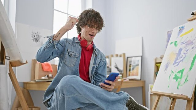 A stylish young man in denim with headphones holds a smartphone in an art studio