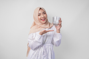 Happy young Asian Muslim woman in a veil hijab pointing to a glass of clear water she holds....