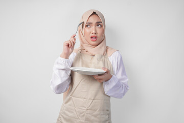 Confused young Asian Muslim woman in hijab and cream apron holding spoon and empty plate with copy...