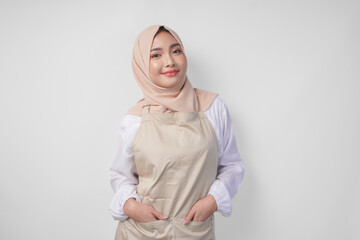 Smiling young Asian Muslim woman in a veil hijab and cream apron doing a formal pose with hands tucked in pockets